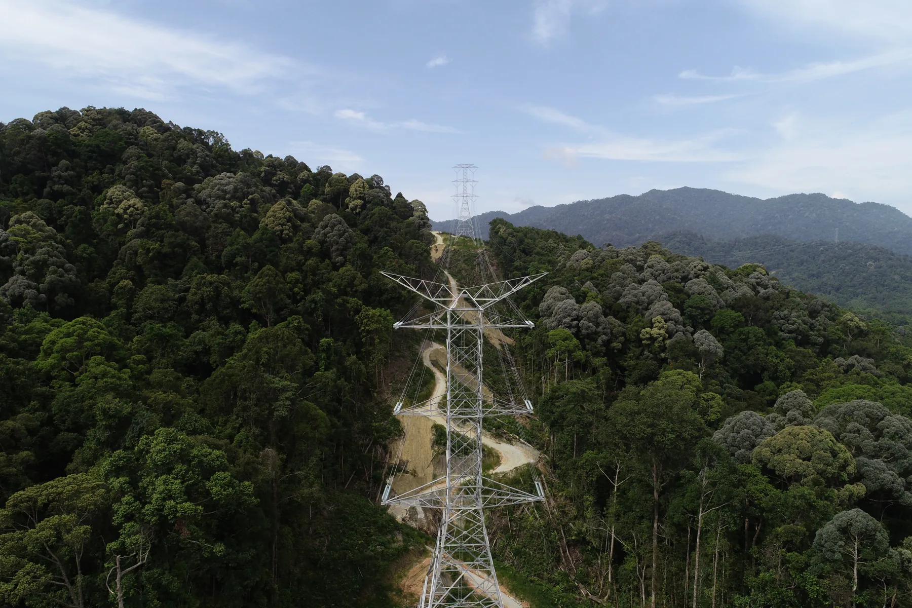 Aerial view of a high-voltage transmission tower on a forested ridge in Malaysia, with the cleared line corridor and access track running back to a second tower