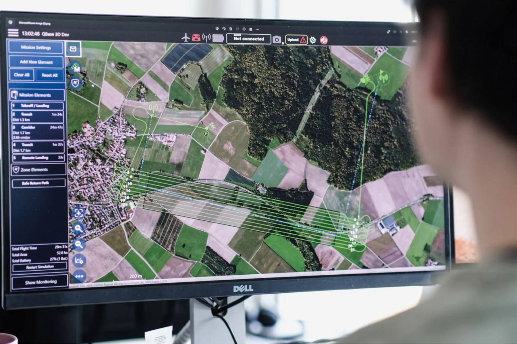 An operator at a widescreen console reviewing a planned drone corridor mission drawn over aerial imagery, with flight time, area and battery totals listed beside it