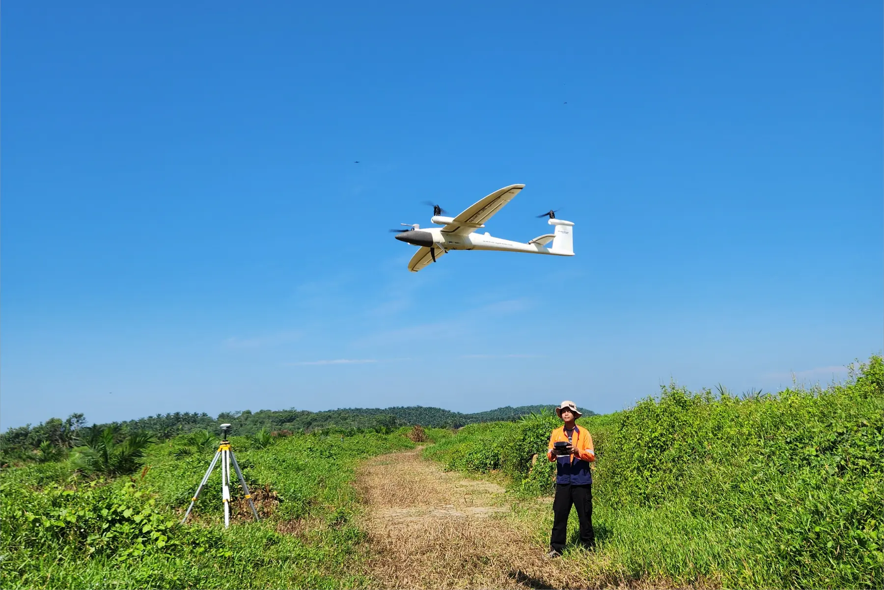 AECA fixed-wing VTOL aircraft flying a mapping run over a plantation track in Malaysia, with an operator at the ground station and a GNSS base station on a tripod below it